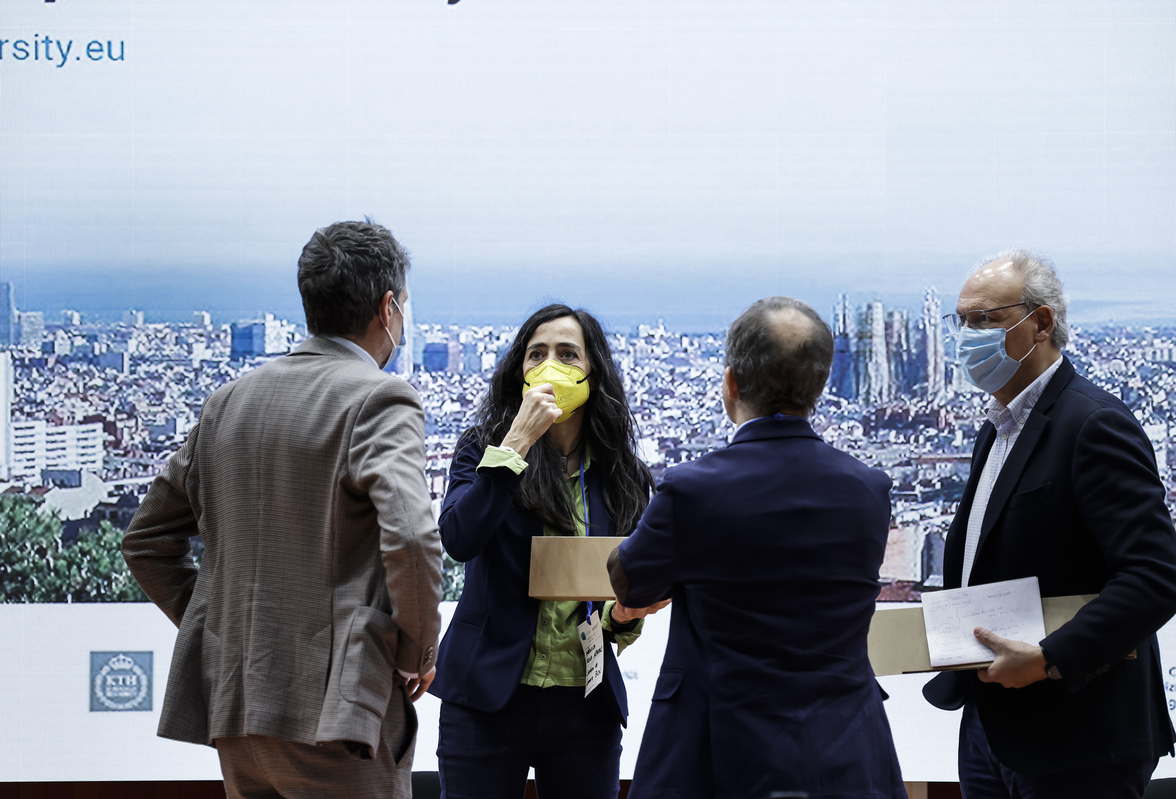 See larger image Attendees of the event discussing with the promotional image of the event screening behind them