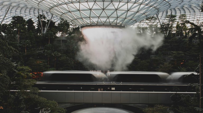 A photograph of a train passing by an interior garden inside of a glass building.