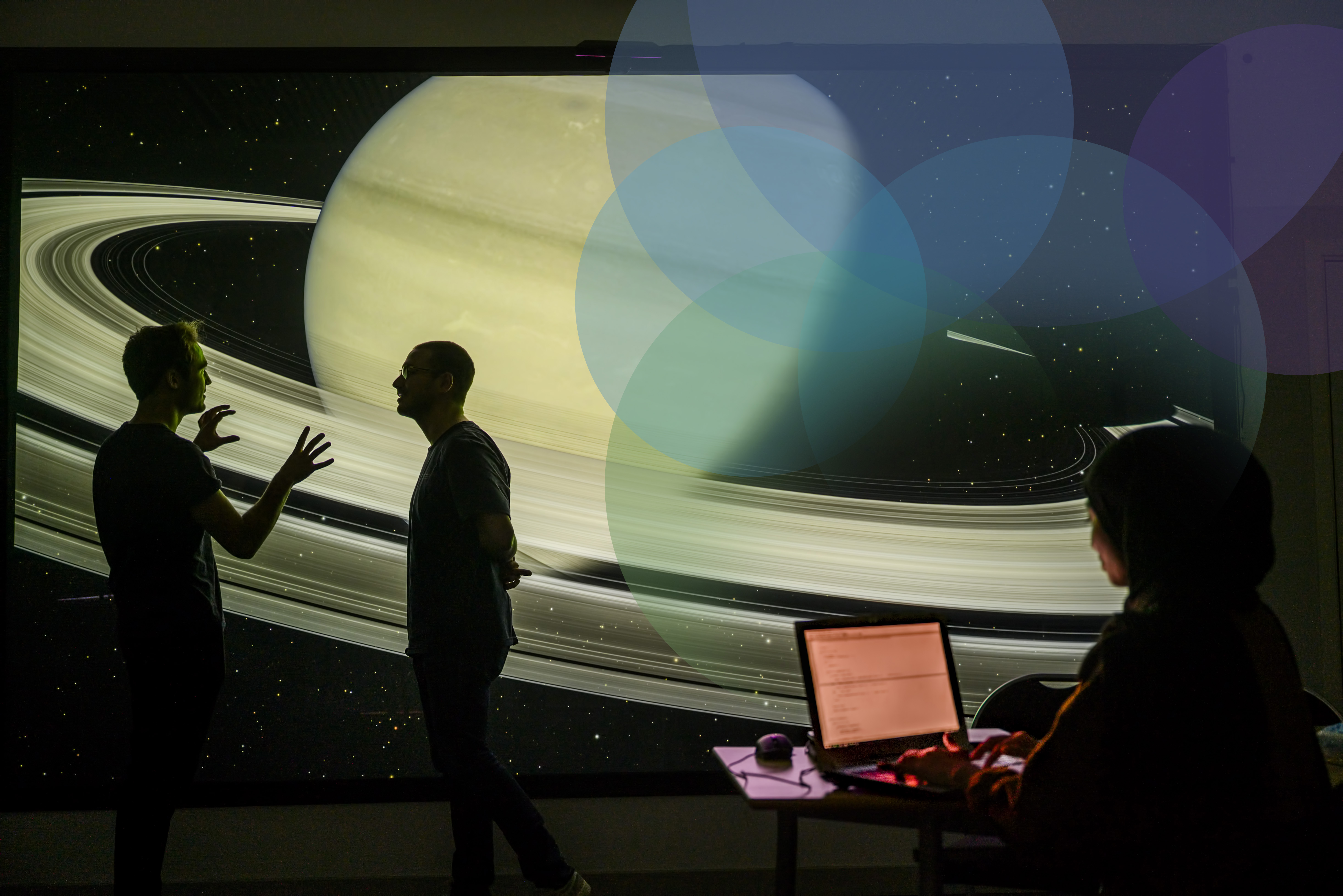 See larger image Two people talking in front of a screen of the planets in space